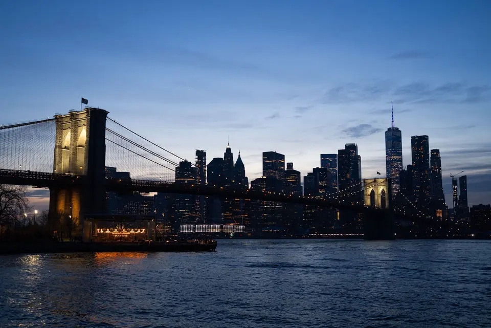 Brooklyn Bridge lit at dusk with the Manhattan skyline behind it, seen across the East River