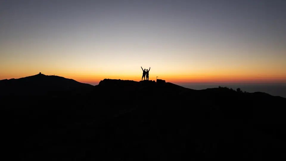 Two silhouetted hikers with arms raised on a mountain ridge at sunset